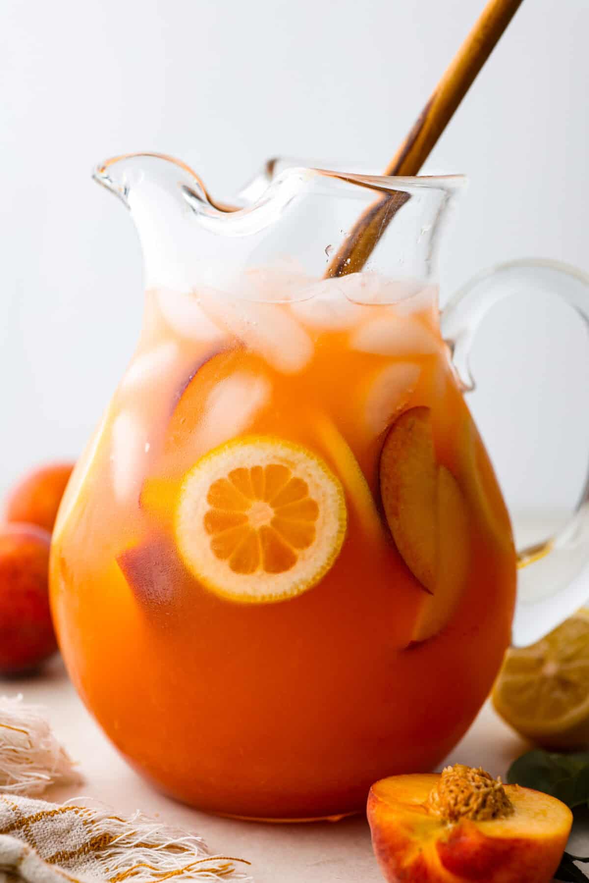 This is the absolute BEST fresh peach lemonade recipe! Sweet juicy peaches combined with tart, tangy lemons make the most refreshing drink. You will want to have a pitcher of this in your fridge throughout the peach season! Side shot of Peach lemonade in a glass pitcher with a wooden spoon sticking.